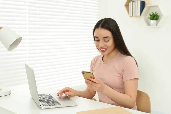 家庭工作场所在房间里 一个快乐的女人 带着智能手机靠近笔记本电脑 坐在白色的书桌上 — 图库照片