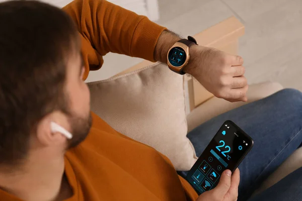 stock image Man setting smart home control system via smartwatch and mobile phone indoors. App interface with icons on display