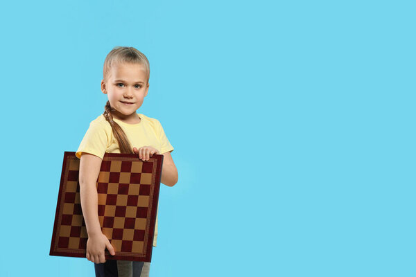 Cute girl holding chessboard on light blue background. Space for text