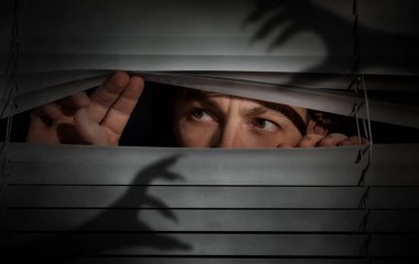 Worried man looking through window blinds into darkness. Shadow of hands with long claws. Paranoia concept clipart
