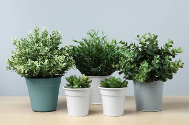 Tahta masadaki saksılarda, açık gri arka planda birçok farklı yapay bitki var.