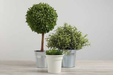 Tahta masadaki saksılarda, açık gri arka planda birçok farklı yapay bitki var.