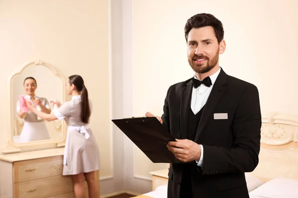 stock image Man wearing suit with clipboard checking maid's work in hotel room, space for text. Professional butler courses