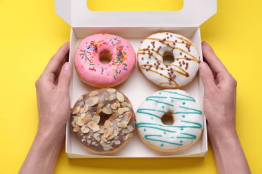 Woman holding box with tasty glazed donuts on yellow background, top view