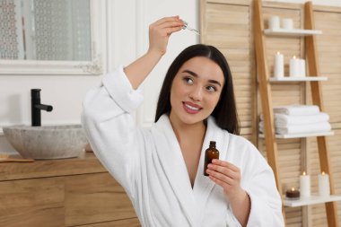 Şişeli mutlu genç kadın banyodaki saç köklerine gerekli yağı sürüyor.
