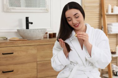 Happy young woman with bottle applying essential oil onto hair in bathroom
