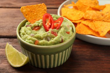 Bowl of delicious guacamole, nachos chips and lime on wooden table, closeup