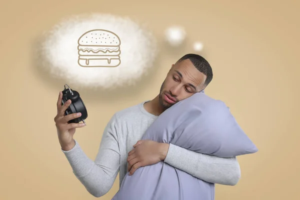 Insomnia. Man with pillow looking at alarm clock on beige background. He can`t fall asleep because of hunger. Thought cloud with illustration of burger