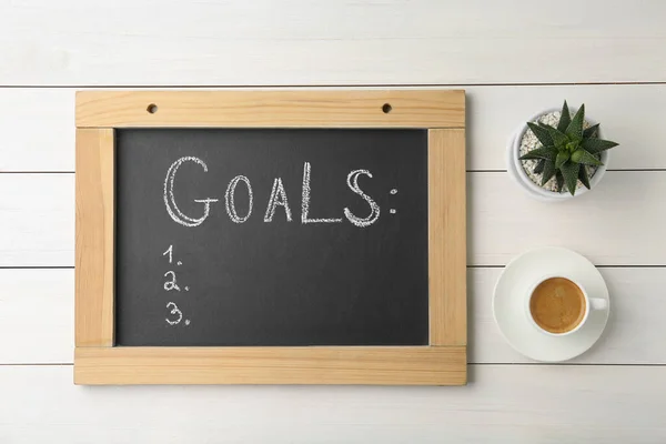 stock image Small blackboard with empty list of goals, houseplant and cup of coffee on white wooden table, flat lay. Space for text