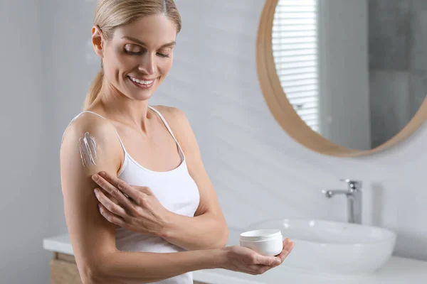 Happy woman applying body cream onto arm in bathroom, space for text