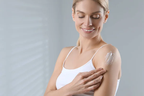 Happy woman applying body cream onto arm indoors, space for text