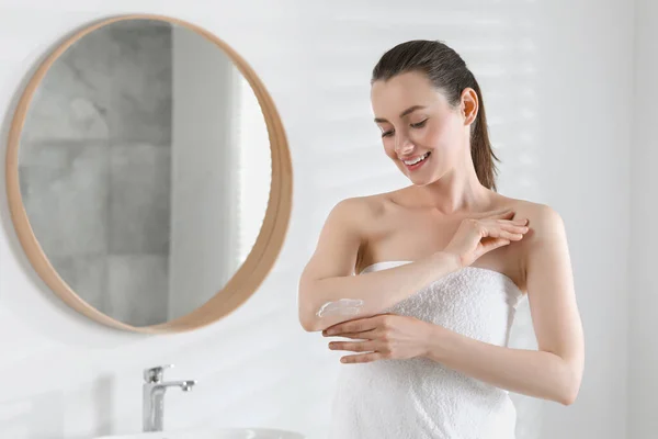 Happy woman applying body cream onto elbow in bathroom, space for text