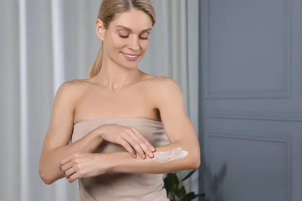 Happy woman applying body cream onto arm indoors, space for text