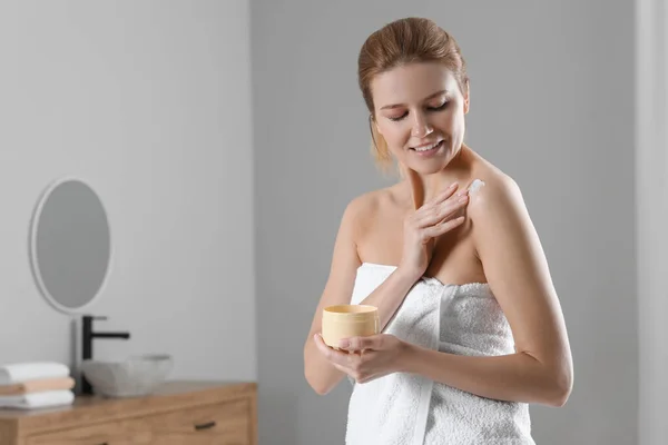 Happy woman applying body cream onto shoulder in bathroom, space for text