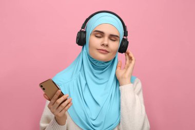 Türbanlı ve kulaklıklı Müslüman bir kadın pembe arka planda akıllı telefonlu müzik dinliyor.