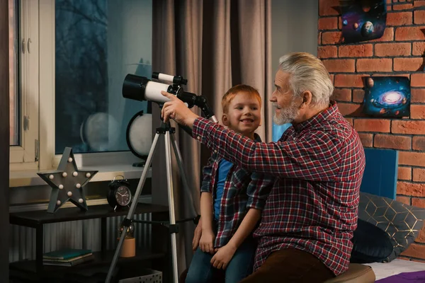 Stock image Little boy with his grandfather using telescope to look at stars in room