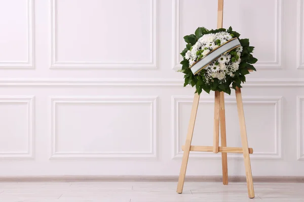 stock image Funeral wreath of flowers with ribbon on wooden stand near white wall indoors. Space for text