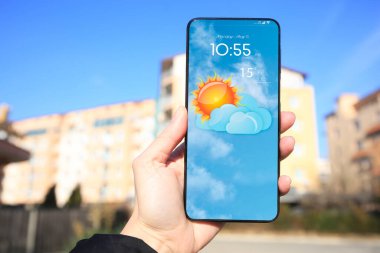 Güneşli bir günde açık hava telefonunda hava durumu uygulaması kullanan bir adam, yakın plan. Ekranda bulutlu güneşin verileri ve çizimleri