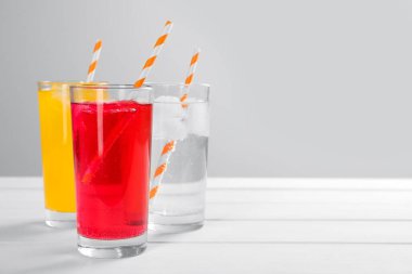 Buzlu farklı tazeleyici soda bardakları ve açık gri arka planda beyaz ahşap masa üzerinde pipetler. Metin için boşluk
