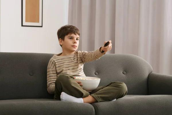 stock image Little boy holding bowl of popcorn and changing TV channels with remote control on sofa at home