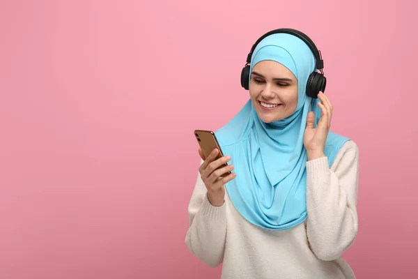 stock image Muslim woman in hijab and headphones listening to music with smartphone on pink background, space for text