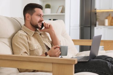 Evdeki kanepede dizüstü bilgisayarla çalışan bir adam telefonda konuşuyor.