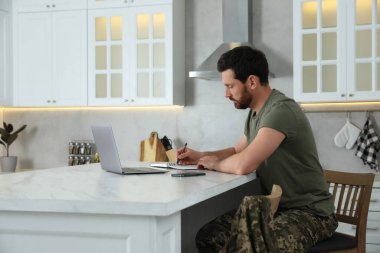 Asker, mutfaktaki beyaz mermer masada dizüstü bilgisayarla çalışırken not alıyor. Askerlik.