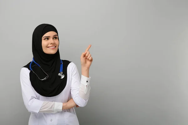 穿着头巾和医疗制服的穆斯林妇女指着浅灰色背景和文字空间的东西 — 图库照片