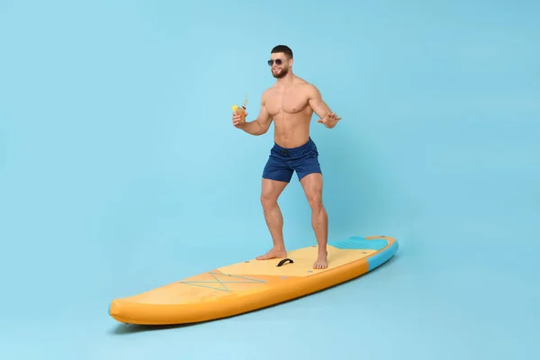 Happy man with refreshing drink balancing on SUP board against light blue background