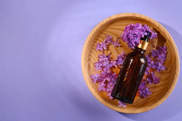 Bottle with essential oil, lilac flowers and water in bowl on violet background, top view. Space for text