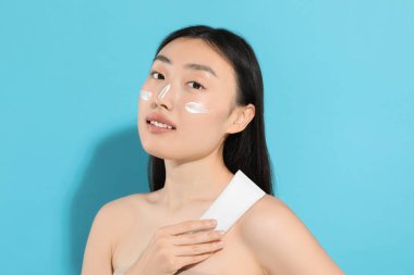 Yüzünde güneş kremi olan güzel genç bir kadın açık mavi arka planda güneş kremi tutuyor.