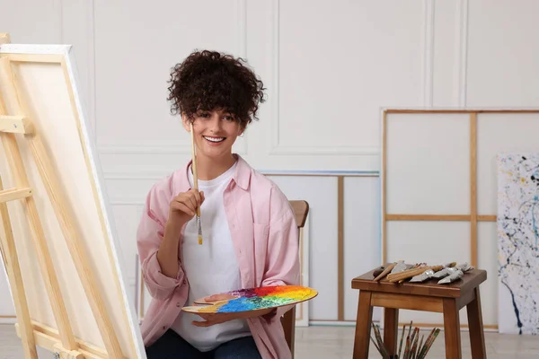 stock image Young woman holding brush and artist`s palette near easel with canvas