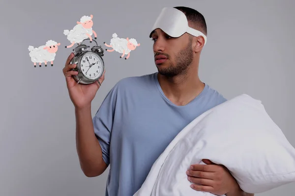 Insomnia. Tired man with pillow checking time on light grey background. Illustrations of sheep jumping over alarm clock