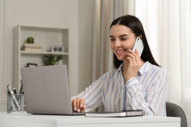 Beyaz masadaki bilgisayarla çalışırken akıllı telefondan konuşan mutlu bir kadın.