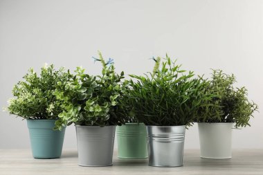 Tahta masadaki saksılarda, açık gri arka planda birçok farklı yapay bitki var.