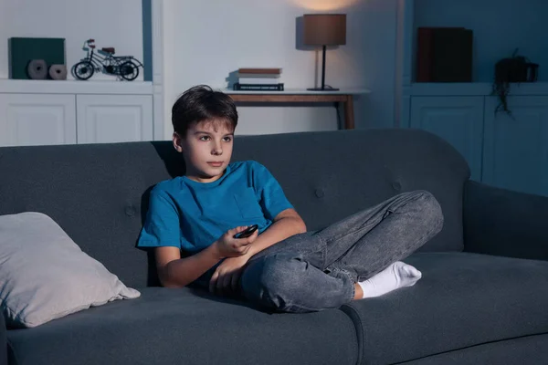 Little boy changing TV channels with remote control on sofa at home