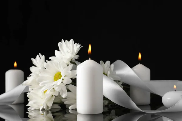 White chrysanthemum flowers and burning candles on black mirror surface in darkness. Funeral symbols