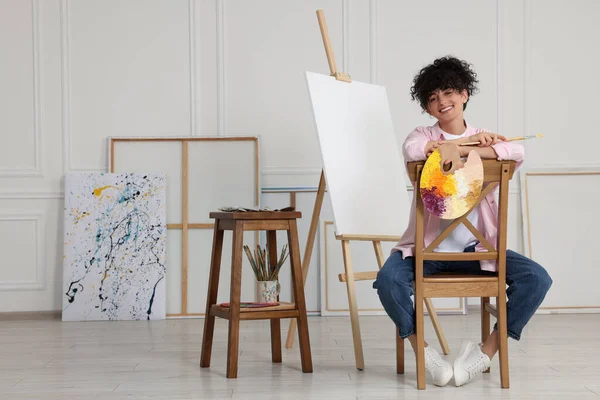 stock image Young woman holding brush and artist`s palette near easel with canvas