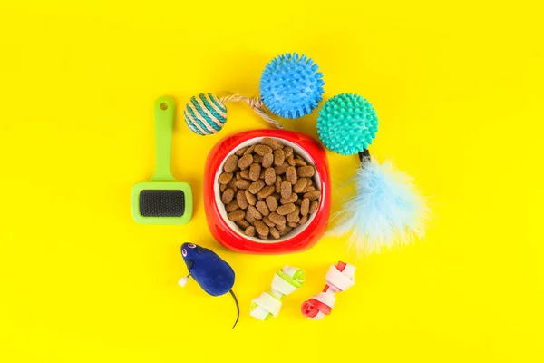 Bowl of dry food, brush and toys on yellow background, flat lay. Pet shop goods