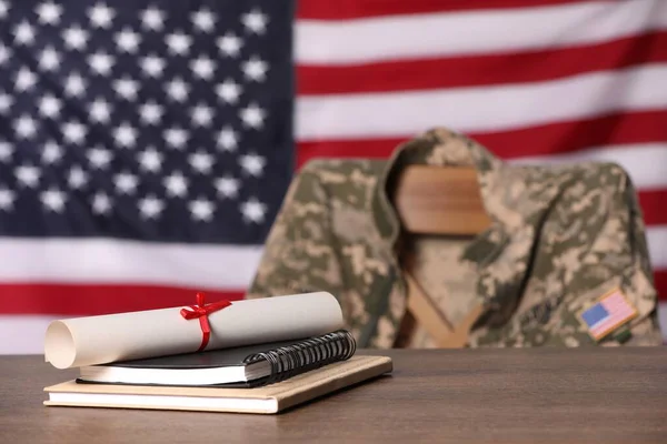 Notebooks and diploma on wooden table, chair with soldier uniform against flag of United States indoors. Military education
