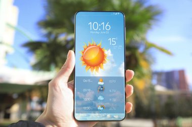 Güneşli bir günde açık hava telefonunda hava durumu uygulaması kullanan bir adam, yakın plan. Veri, güneş ve diğer illüstrasyonlar ekranda