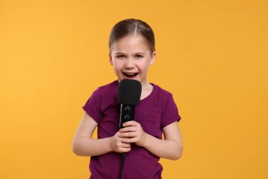 Sarı arka planda mikrofonlu şirin küçük kız şarkı söylüyor.
