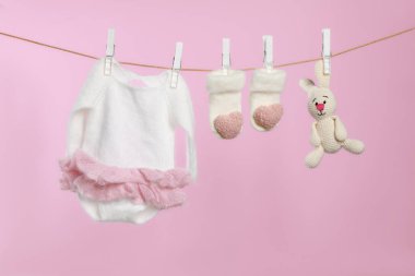 Bebek kıyafetleri ve el yapımı oyuncak pembe arka planda çamaşır ipinde kuruyor.