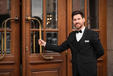 Butler in elegant suit opening wooden hotel door. Space for text clipart