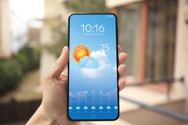 Kadın hava durumunu kontrol ediyor. Açık havada akıllı telefon uygulaması kullanıyor. Yakın plan. Ekranda bulutlu güneşin verileri ve çizimleri