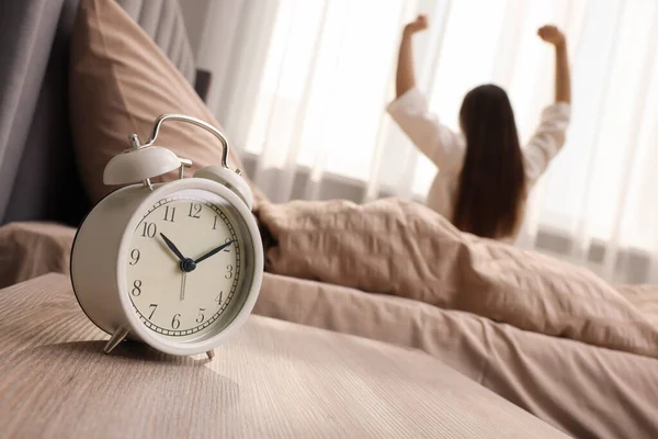 It's lazy morning o'clock. Alarm clock on bedside table and woman stretching in room, selective focus