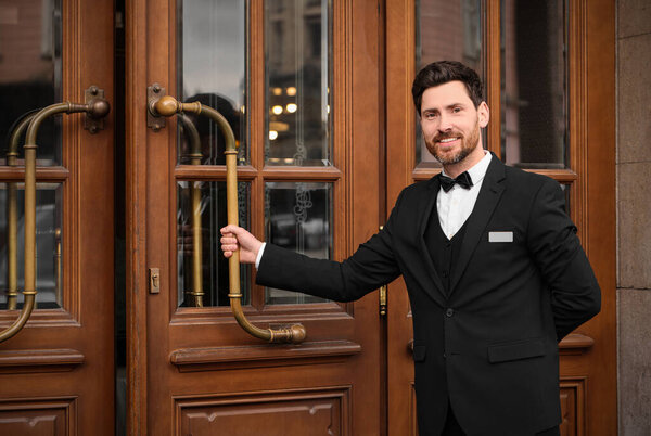 Butler in elegant suit opening wooden hotel door. Space for text