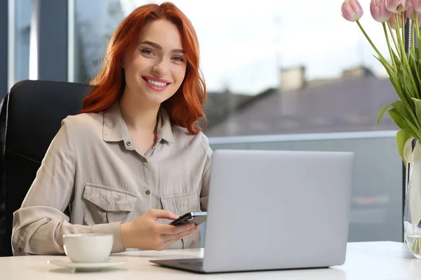 一个快乐的女人 在办公室的白色办公桌前 用智能手机在笔记本电脑上工作 — 图库照片