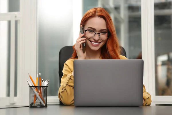 快乐的女人一边在办公室里用笔记本电脑一边用智能手机聊天 — 图库照片
