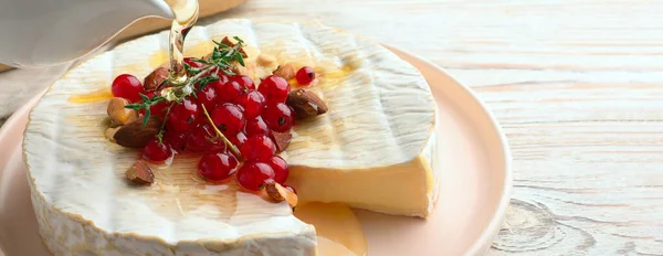 Pouring honey onto tasty brie cheese with red currant and almonds on white wooden table, closeup. Banner design with space for text